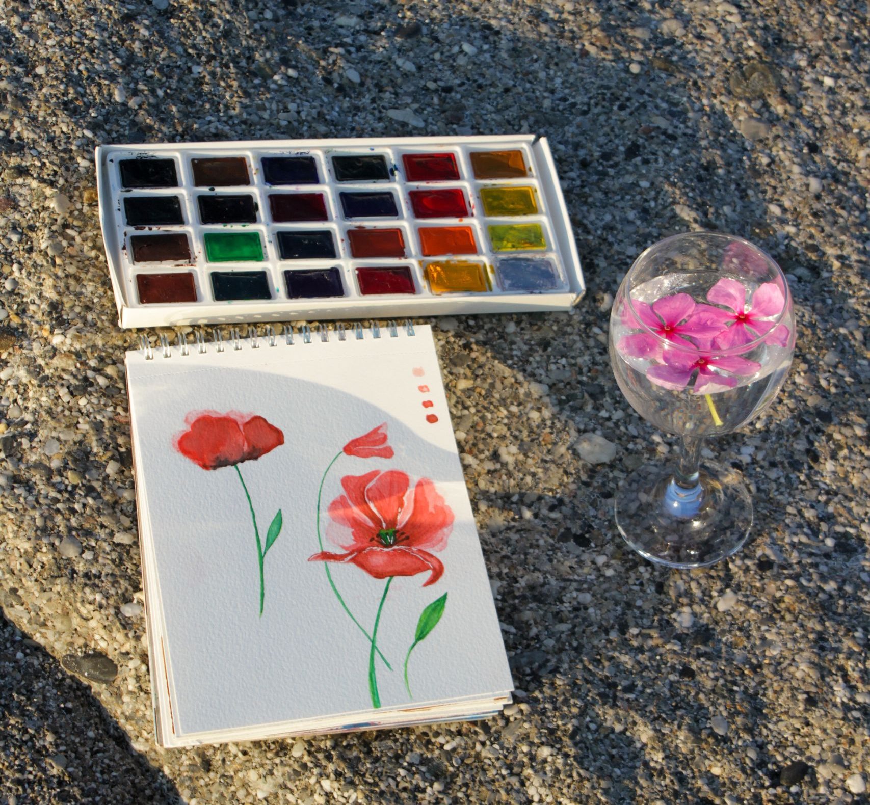 Watercolor painting set on a sandy beach with a painted flower notebook and petals in a glass.